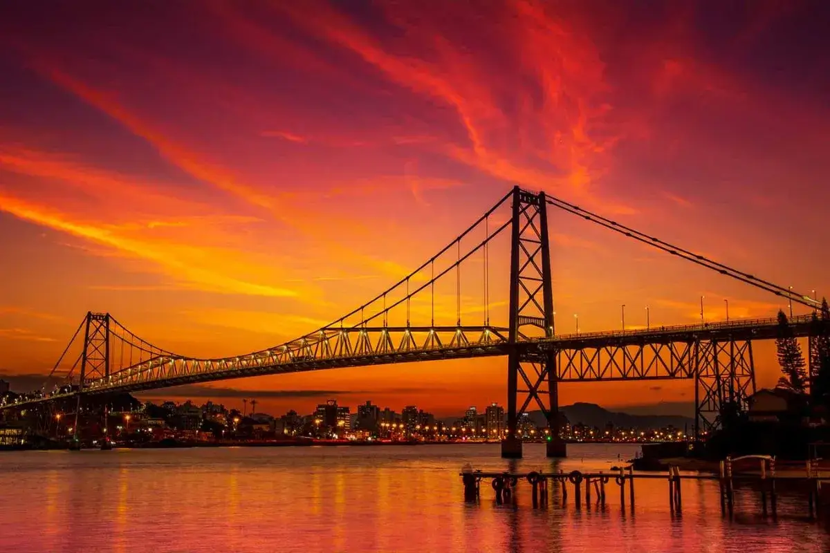 Vista da Ponte Hercílio Luz em Florianópolis, Brasil, sobre a Baía Norte, com a estrutura metálica da ponte em destaque e a cidade ao fundo, sob um céu alaranjado ao pôr do sol refletido na água.