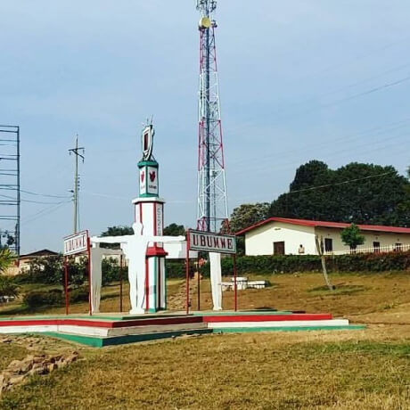 Monumento localizado em uma área aberta de Muramvya, Burundi, com uma estrutura vertical decorada nas cores da bandeira nacional, cercada por um pequeno jardim e edificações simples ao fundo, sob céu azul.