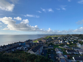 Vista panorâmica de uma cidade costeira com casas alinhadas, ruas residenciais e o mar ao fundo sob céu azul com nuvens.