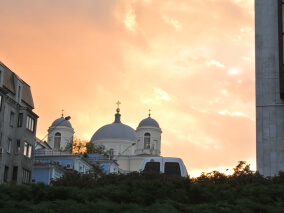 Igreja histórica com cúpulas claras ao fundo, vista entre prédios urbanos durante o pôr do sol.