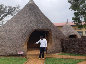 Homem em frente a uma construção tradicional com telhado de palha, em área cultural ao ar livre.